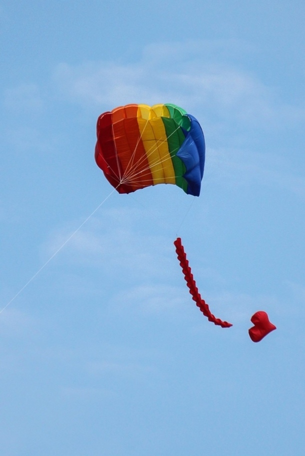 Ein Farbbild im Hochformat. Ein Flugdrachen viereckig in Regenbogenfarben mit einem Schweif an dessen Ende ein Herz hängt.